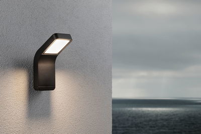 Schwarze LED-Außenwandleuchte aus Metall mit warmweißem Licht an grauer Wand mit Meerblick