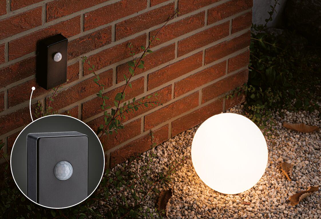 Black Zigbee motion sensor with IP44 protection mounted on red brick wall outdoors near glowing sphere light.