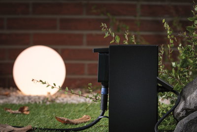 Boîtier extérieur noir avec câble devant une lampe sphérique éclairée dans un jardin avec herbe et pierre