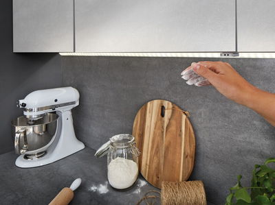 Køkkenbord med hvid røremaskine, træbræt, glas med mel og gråt LED-underkabinetlys.