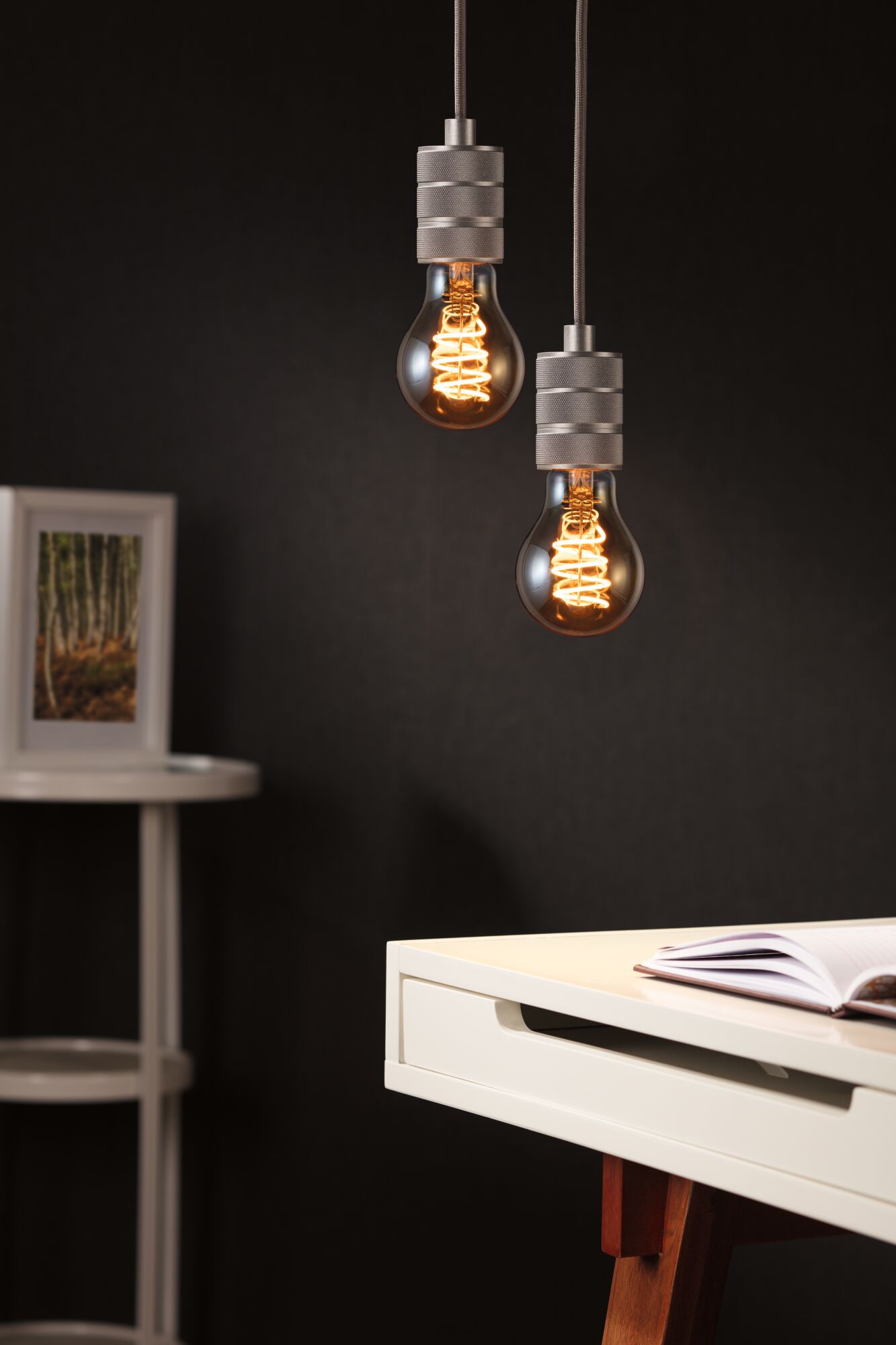 Zwei hängende Glühbirnen mit warmem Licht und grauen Fassungen vor dunklem Hintergrund im modernen Arbeitszimmer.