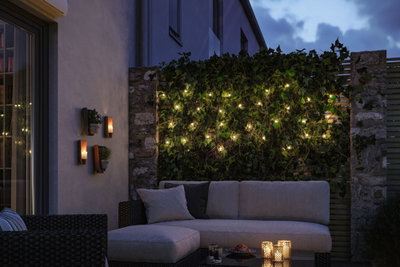 Hyggelig terrasse med grå polstret sofa, grøn plantevæg og varm hvide LED-lyskæder.