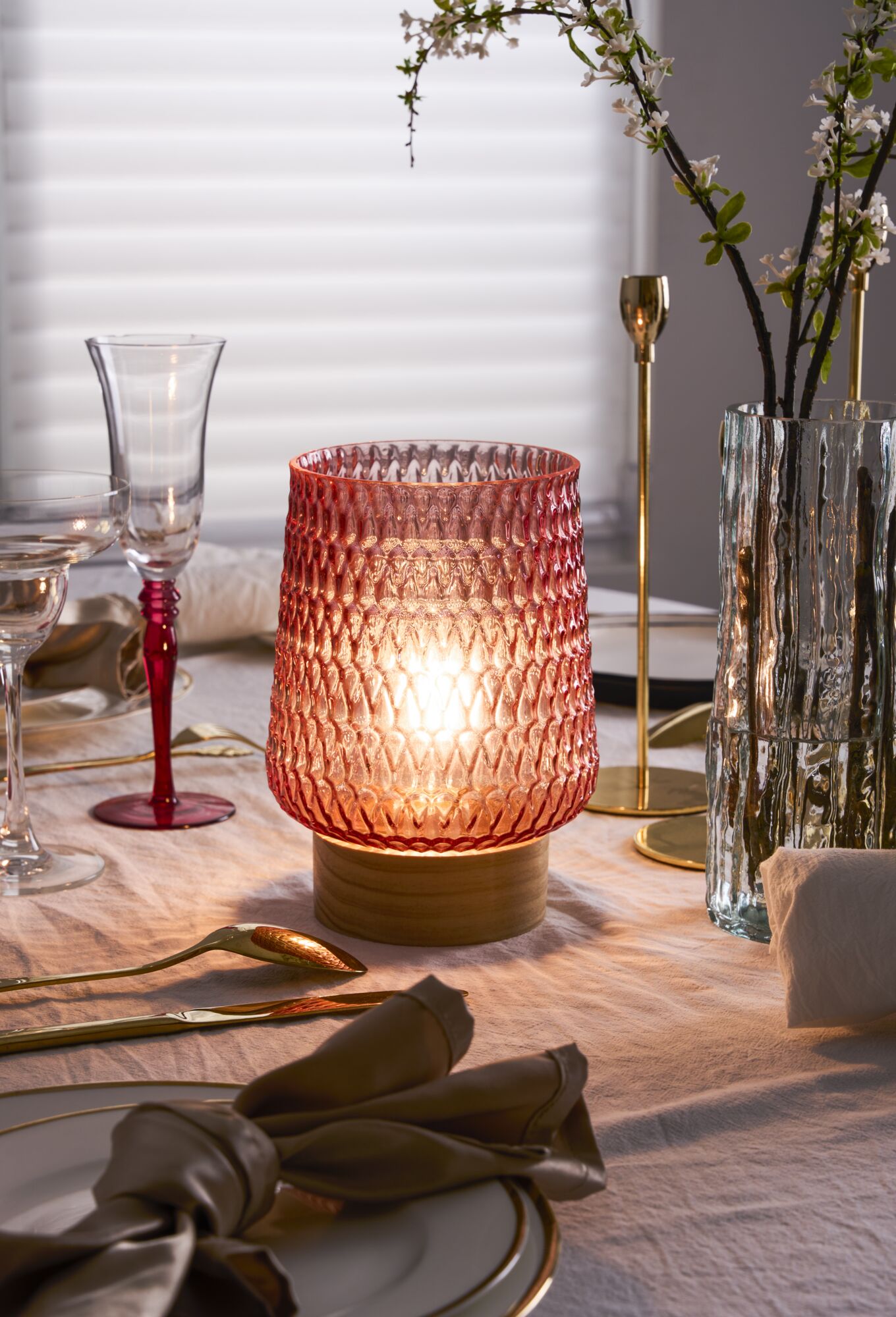Rosa Glas-Tischleuchte mit Holzfuß auf gedecktem Esstisch mit goldfarbenem Besteck und Glaswaren