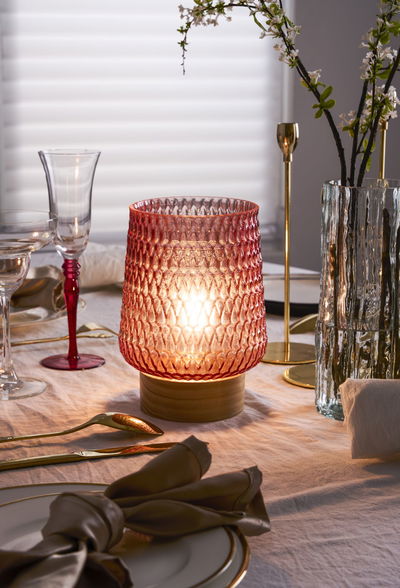 Rosa Glas-Tischleuchte mit Holzfuß auf gedecktem Esstisch mit goldfarbenem Besteck und Glaswaren