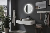 Modern bathroom with round LED mirror, grey wall, white sink, and wooden stool with towels