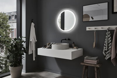 Modern bathroom with round LED mirror, grey wall, white sink, and wooden stool with towels