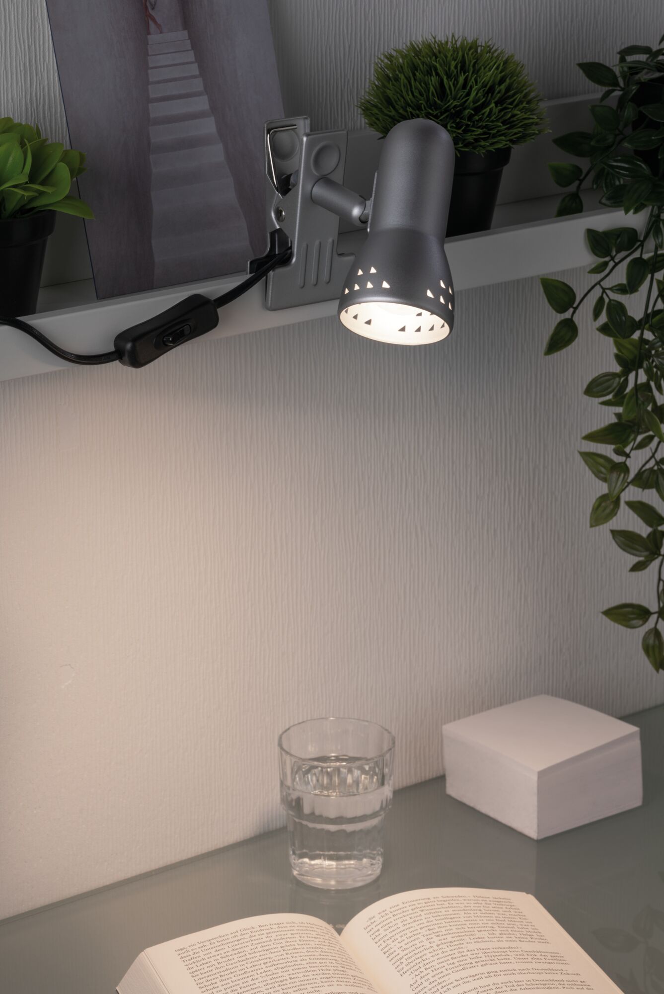 Spot à pince en métal gris avec lumière blanche chaude sur étagère au-dessus d'un bureau avec livre et verre d'eau