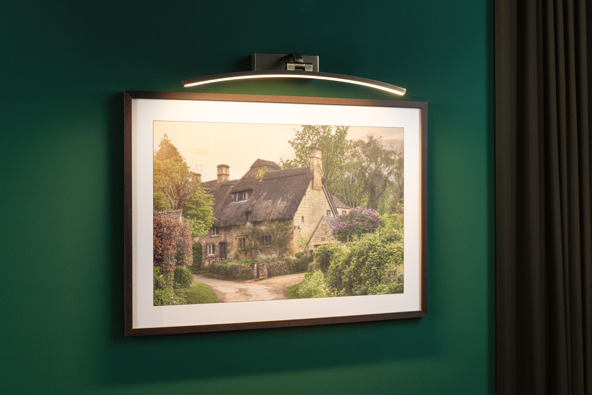 Curved LED picture light in brushed metal above framed countryside house photo on green wall.