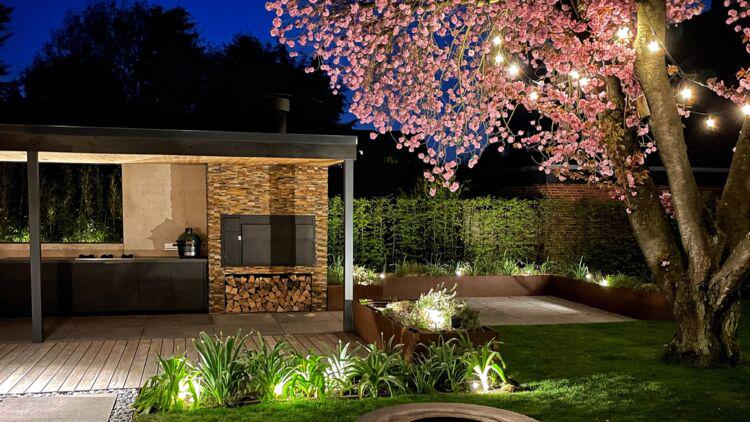 Jardin éclairé avec cuisine extérieure moderne et guirlandes LED blanc chaud sur arbre en fleurs la nuit