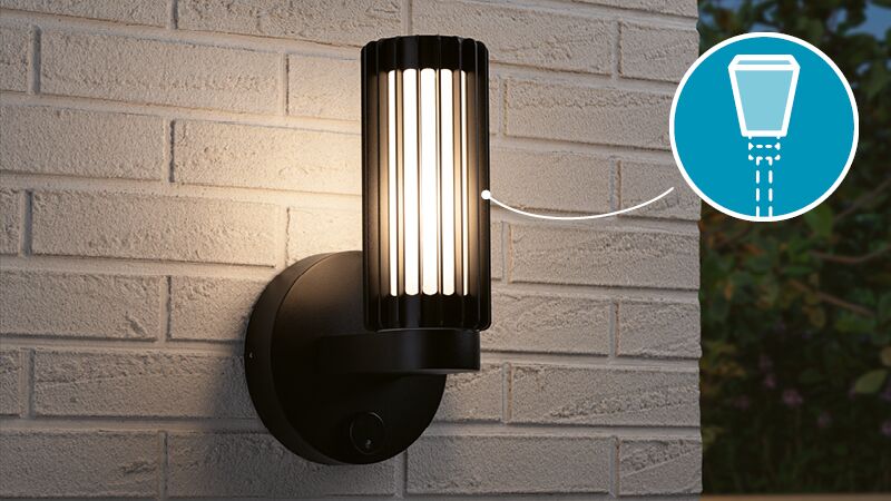 Black metal outdoor wall light with vertical slats and warm white illumination on light brick wall