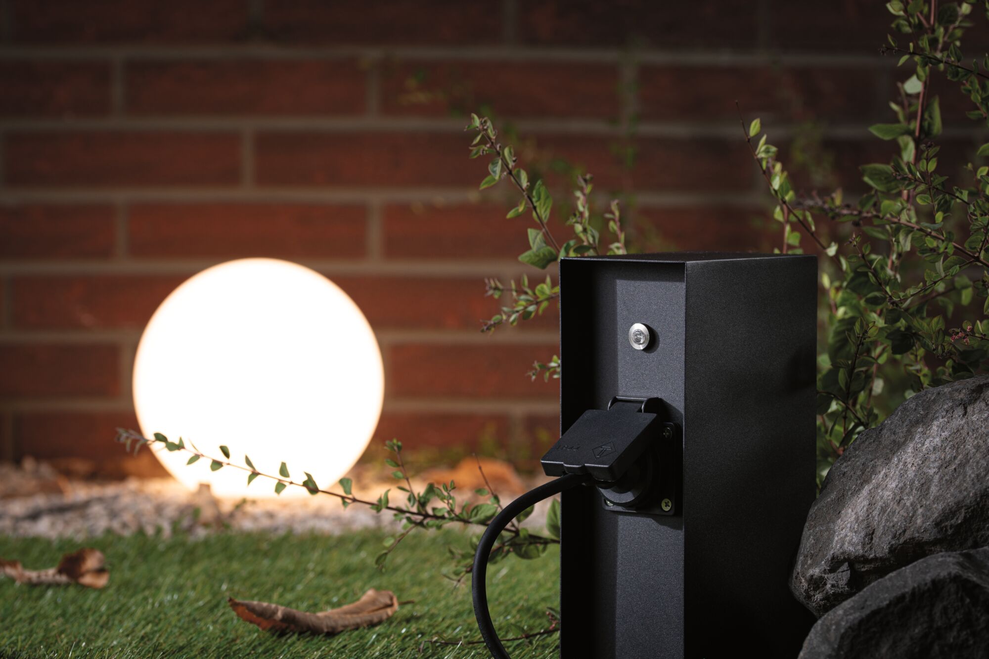 Prise extérieure noire en métal avec câble devant sphère de jardin lumineuse et mur en briques