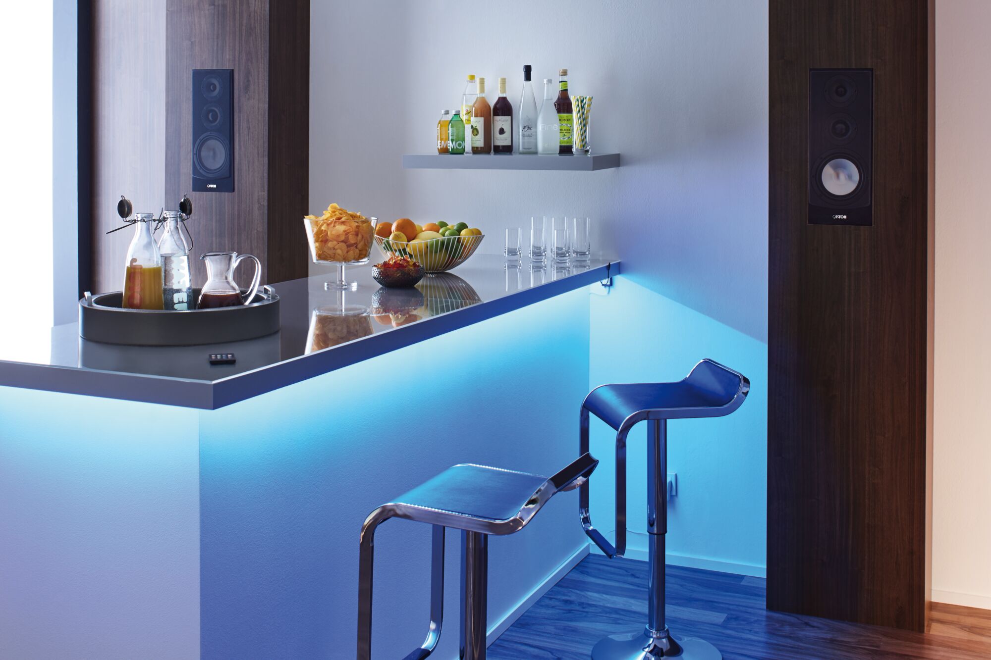Moderne bar med blå LED-belysning, to blå barstole og drikkevarer på en hvid bordplade i en lounge.