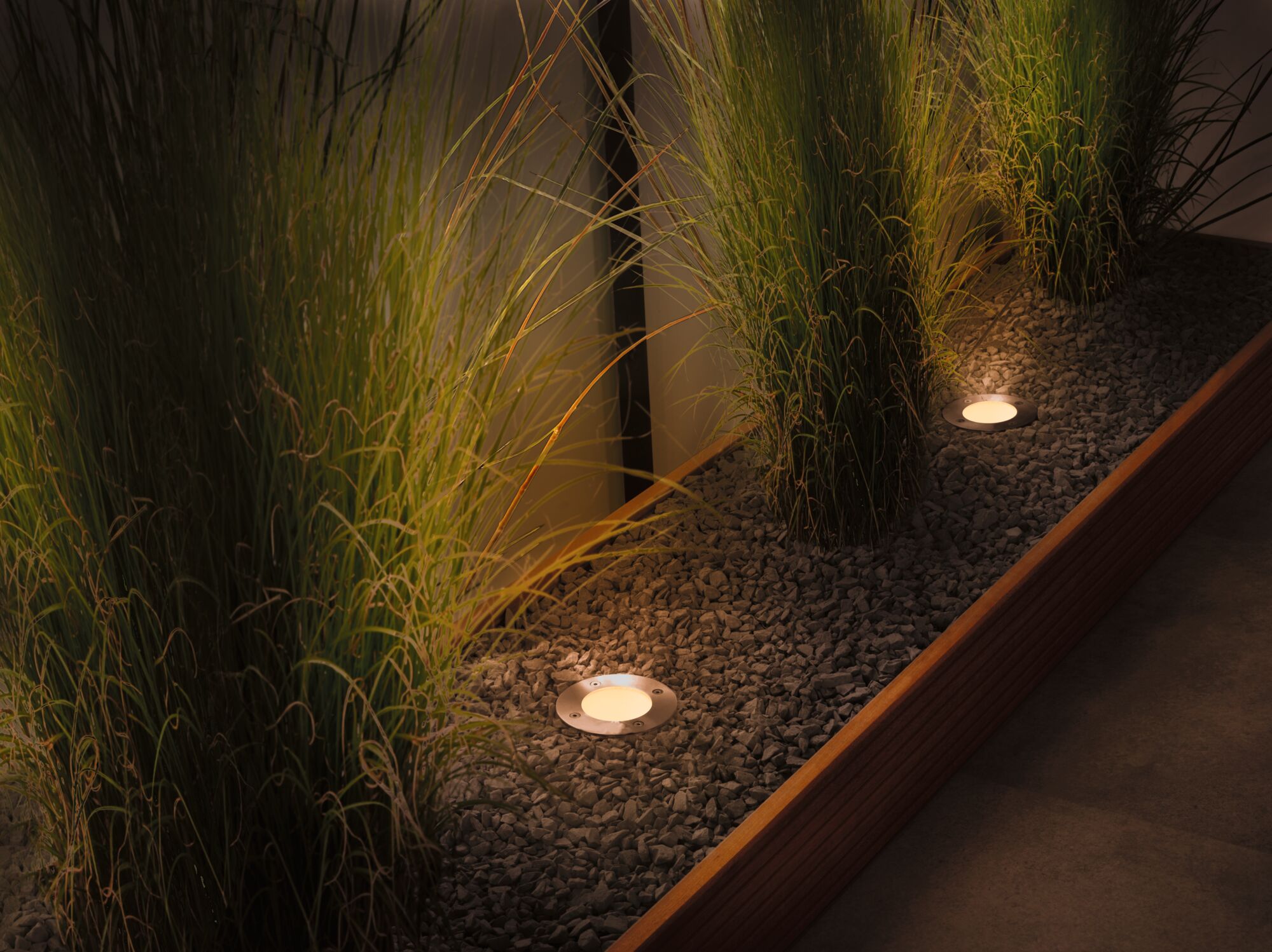 Eingelassene LED-Bodenleuchten aus Edelstahl beleuchten Kiesbeet mit hohen grünen Gräsern im Garten.