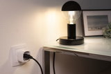 Schwarze Tischlampe mit halbkugelförmiger Glühbirne auf Glasplatte neben weißer Steckdose mit schwarzem Kabel