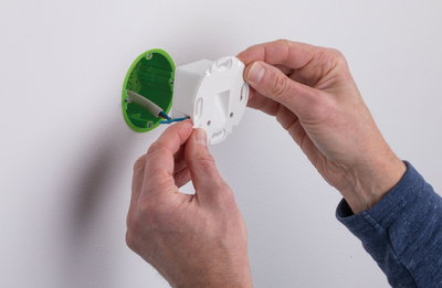 Hands installing white plastic electrical junction box with green insert on white wall for lighting setup