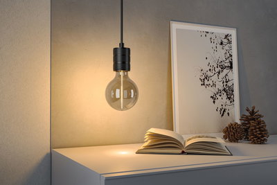 Hanging light bulb with black metal socket illuminating open book on white table with decor items.