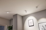 Three white metal LED ceiling spotlights illuminate modern framed pictures in a bright room with plants.