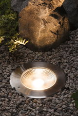 Brushed stainless steel ground spotlight with warm white LED light surrounded by gravel and rocks outdoors