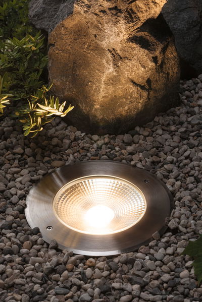 Brushed stainless steel ground spotlight with warm white LED light surrounded by gravel and rocks outdoors