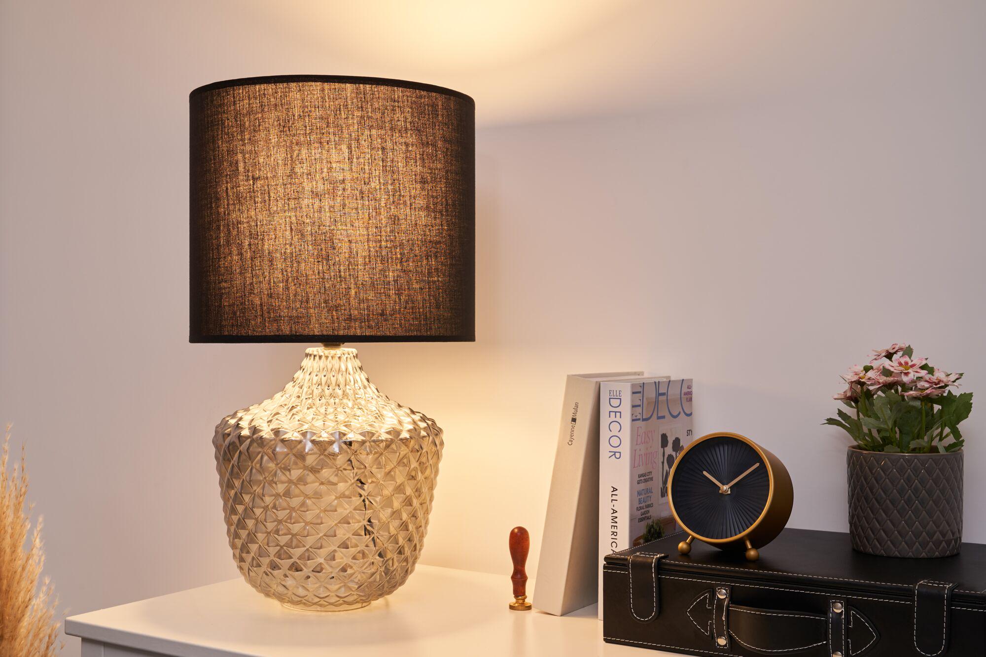 Table lamp with brown fabric shade and textured clear glass base on white table with clock and plant