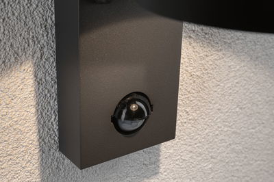 Détecteur de mouvement noir fixé sur mur blanc texturé pour éclairage extérieur et sécurité.