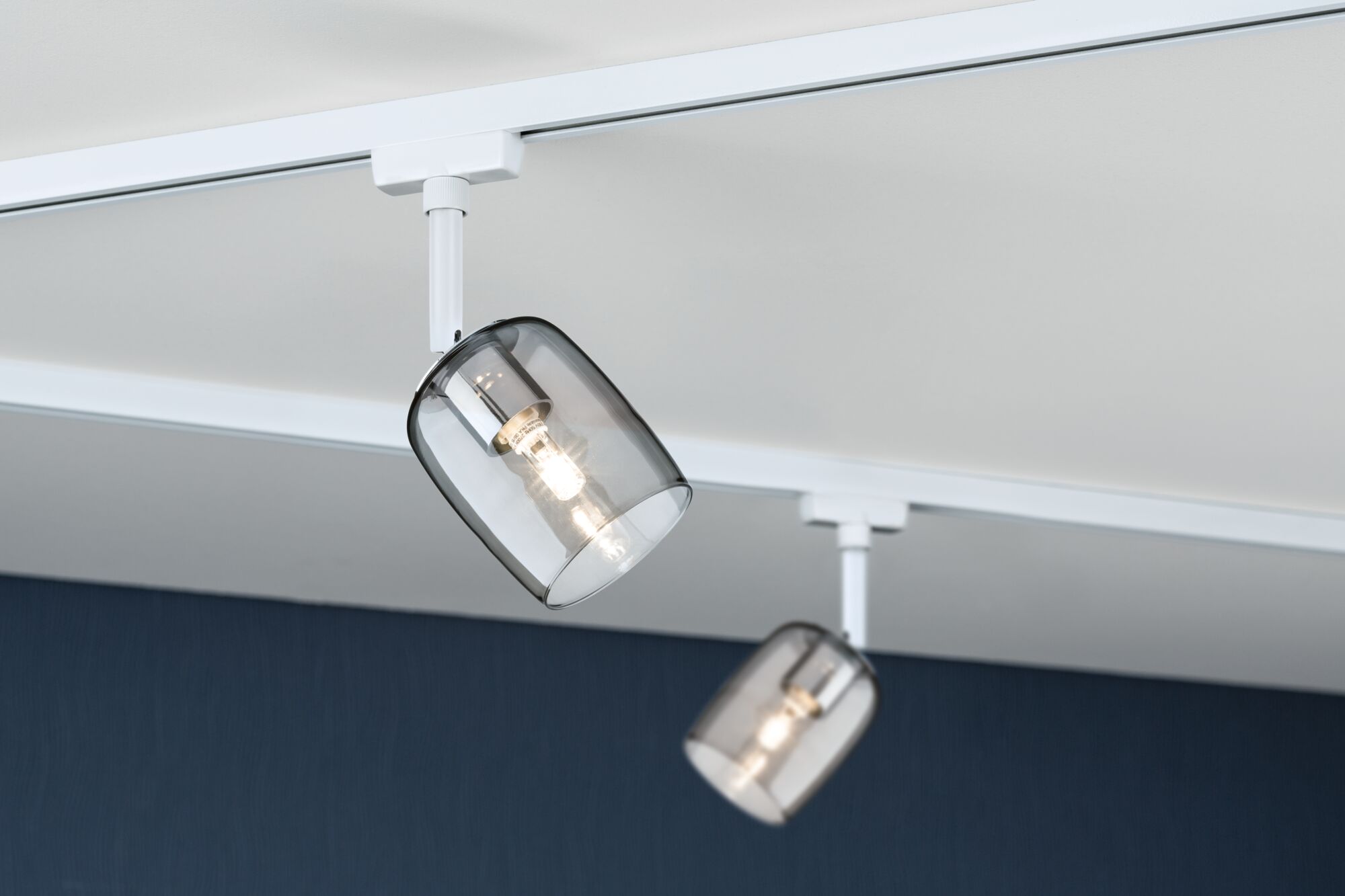 Zwei weiße Schienenspot-Leuchten mit rauchgrauen Glaslampenschirmen an Decke montiert für Wohnraumbeleuchtung.
