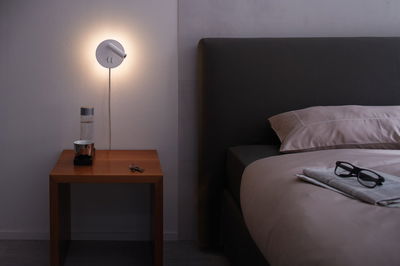 Weiße Wandlampe aus Metall mit warmem Licht über Nachttisch neben dunklem Bett und Brille auf Bettdecke