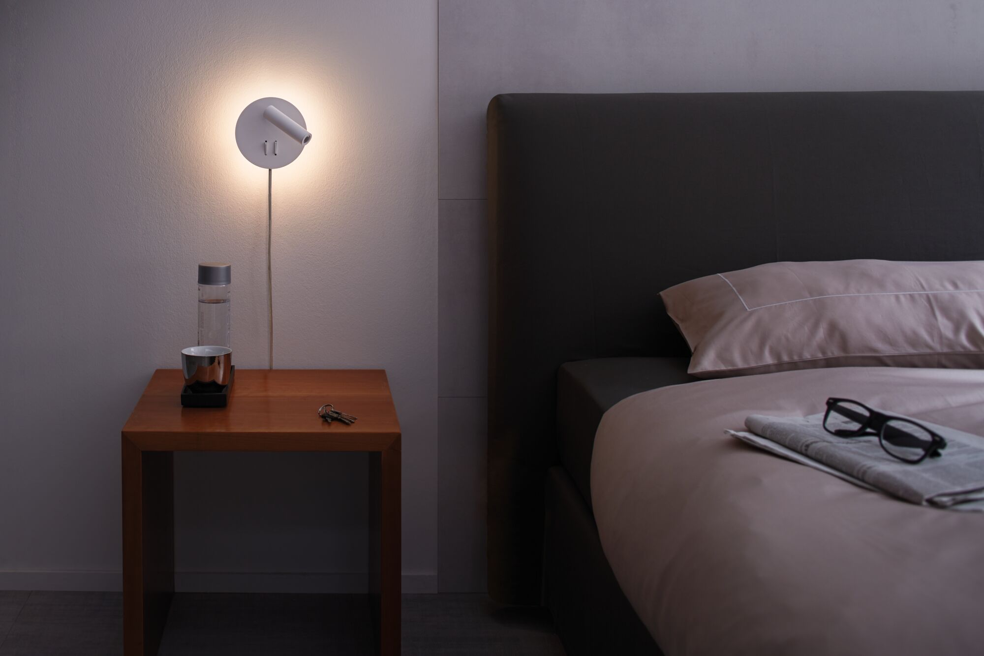 White metal wall lamp with warm light above wooden bedside table next to dark bed with glasses on duvet