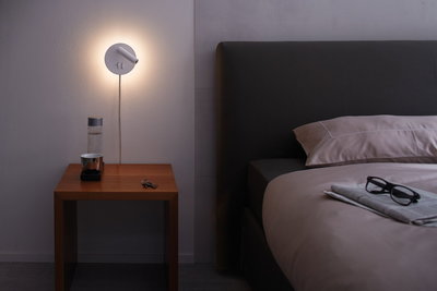 White metal wall lamp with warm light above wooden bedside table next to dark bed with glasses on duvet