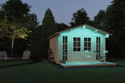 Maison de jardin en bois avec grande terrasse et table en métal blanc, éclairée par lumière LED verte.