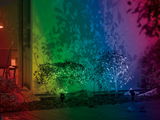 Multicolor LED garden spotlights illuminate green bushes creating colorful shadows on the wall.