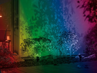 Mehrfarbige LED-Gartenstrahler beleuchten grüne Büsche und erzeugen bunte Schatten an der Hauswand.