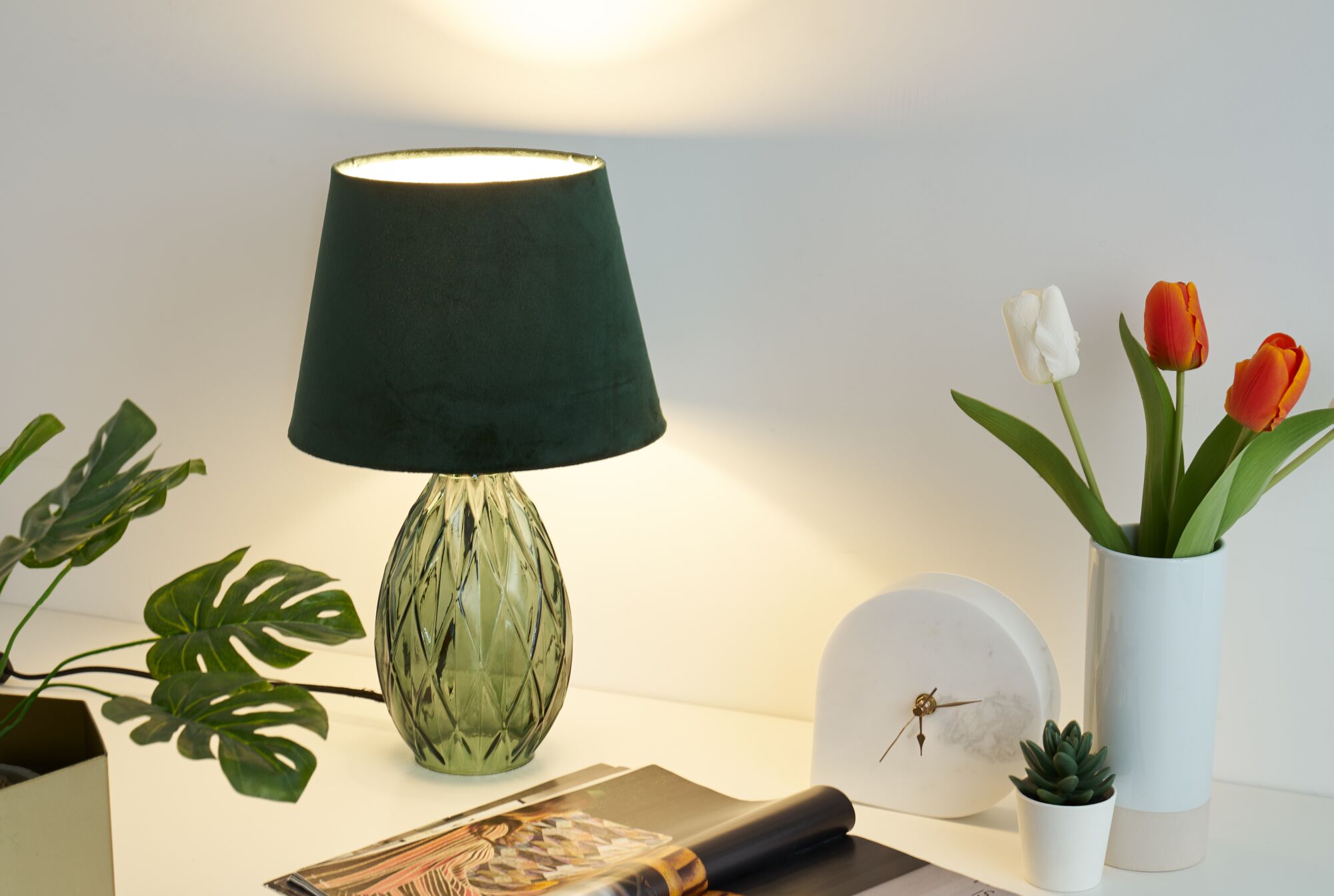 Groene tafellamp met gestructureerde glazen voet en donkergroene stoffen kap op wit bureau met decoratie.