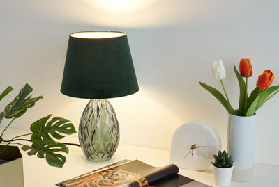 Green glass table lamp with dark green fabric shade beside tulips and white clock on desk