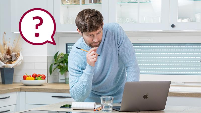 Mann in blauem Pullover arbeitet am Laptop in moderner Küche mit Notizblock und Glas Wasser.