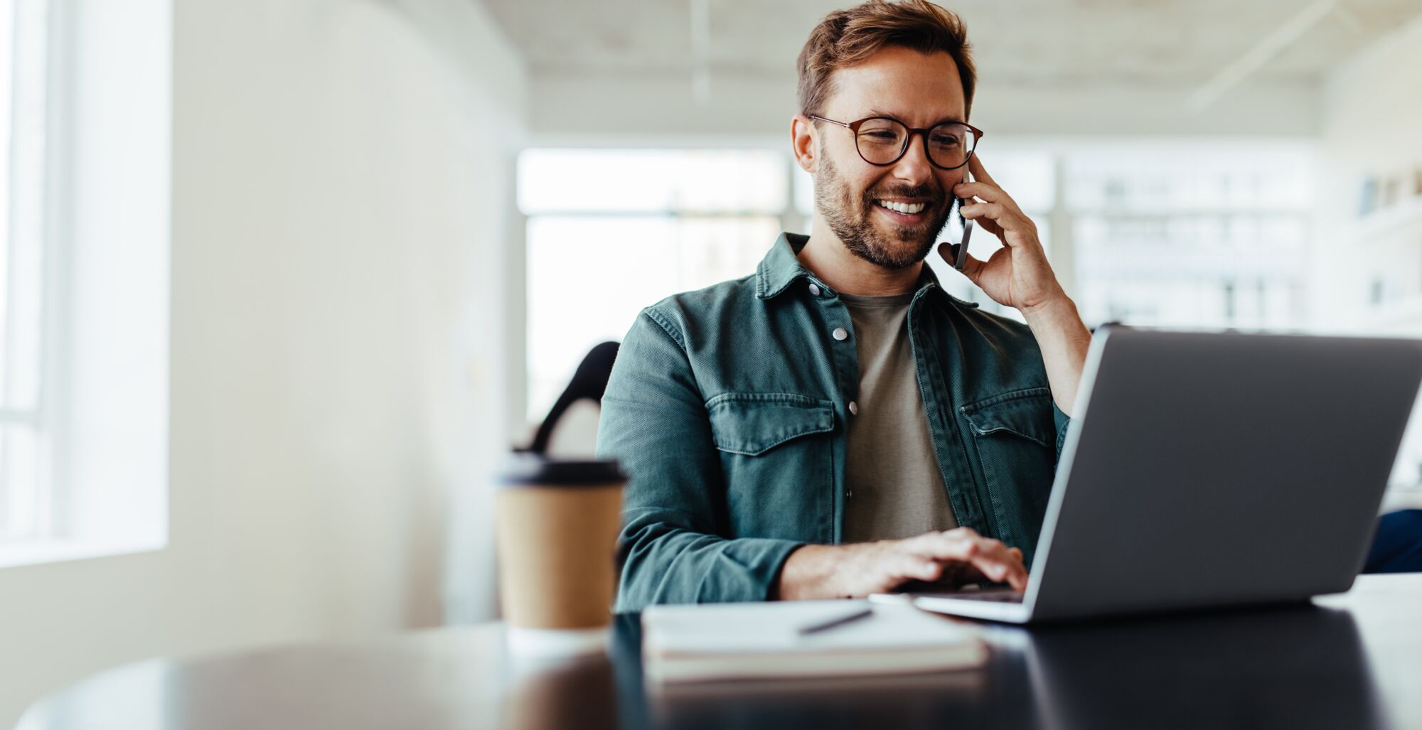 Man with glasses smiling on phone while using laptop in bright office with coffee cup and notebook