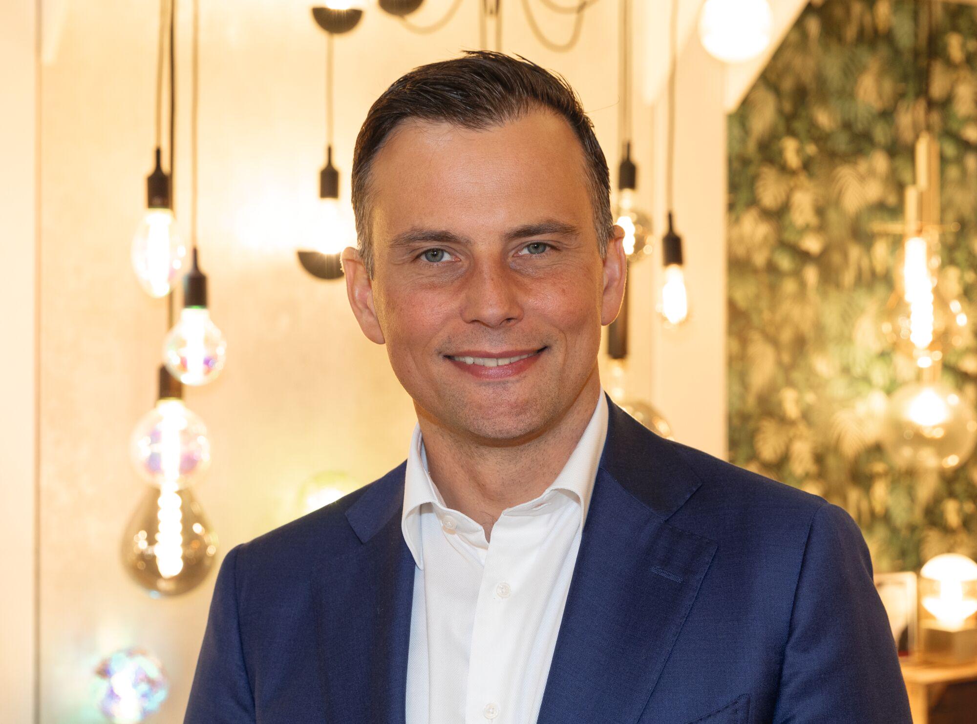 Porträt eines Mannes in blauem Anzug vor warm beleuchteter Wand mit dekorativen Glühbirnen