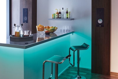 Bar moderne avec éclairage LED turquoise, deux tabourets verts et enceintes murales en bois dans une pièce élégante.
