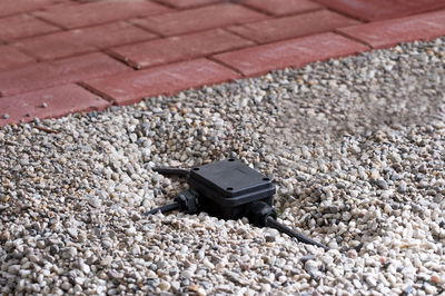 Black plastic junction box with cables on gravel near red paving for outdoor lighting setup