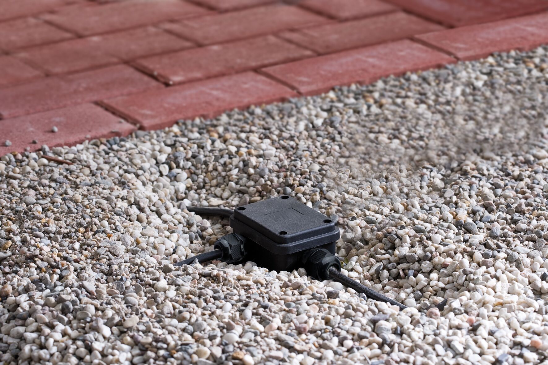 Black plastic junction box with cables on gravel near red paving for outdoor lighting setup