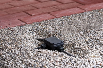 Black plastic junction box with cables in gravel bed next to red paving for outdoor lighting setup