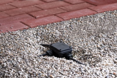 Black plastic junction box with cables in gravel bed next to red paving for outdoor lighting setup