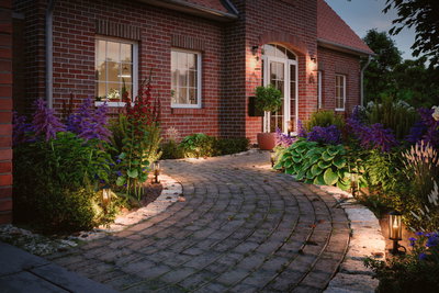 Allée de jardin éclairée avec pavés et lanternes devant une maison en briques rouges avec plantes et fleurs.