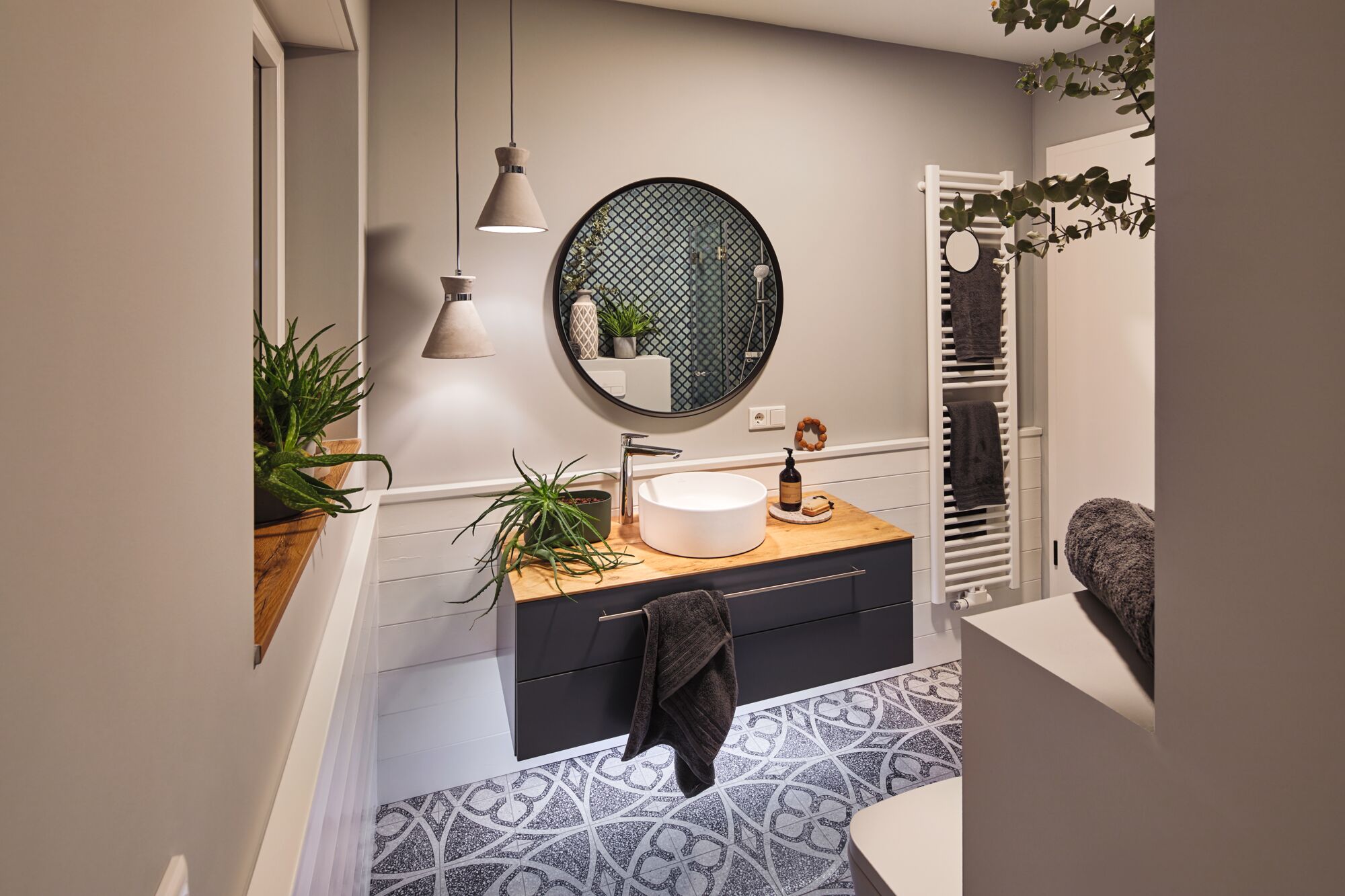 Modern bathroom with dark grey floating vanity, wooden countertop, round mirror, plants, and pendant lights